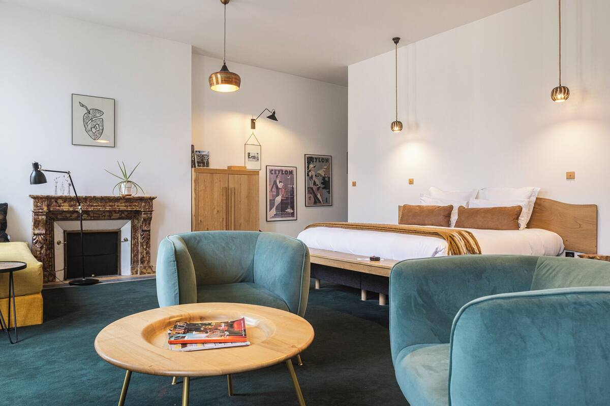 A modern hotel room with a bed, two armchairs, a small wooden table, a fireplace, and artwork on the walls.