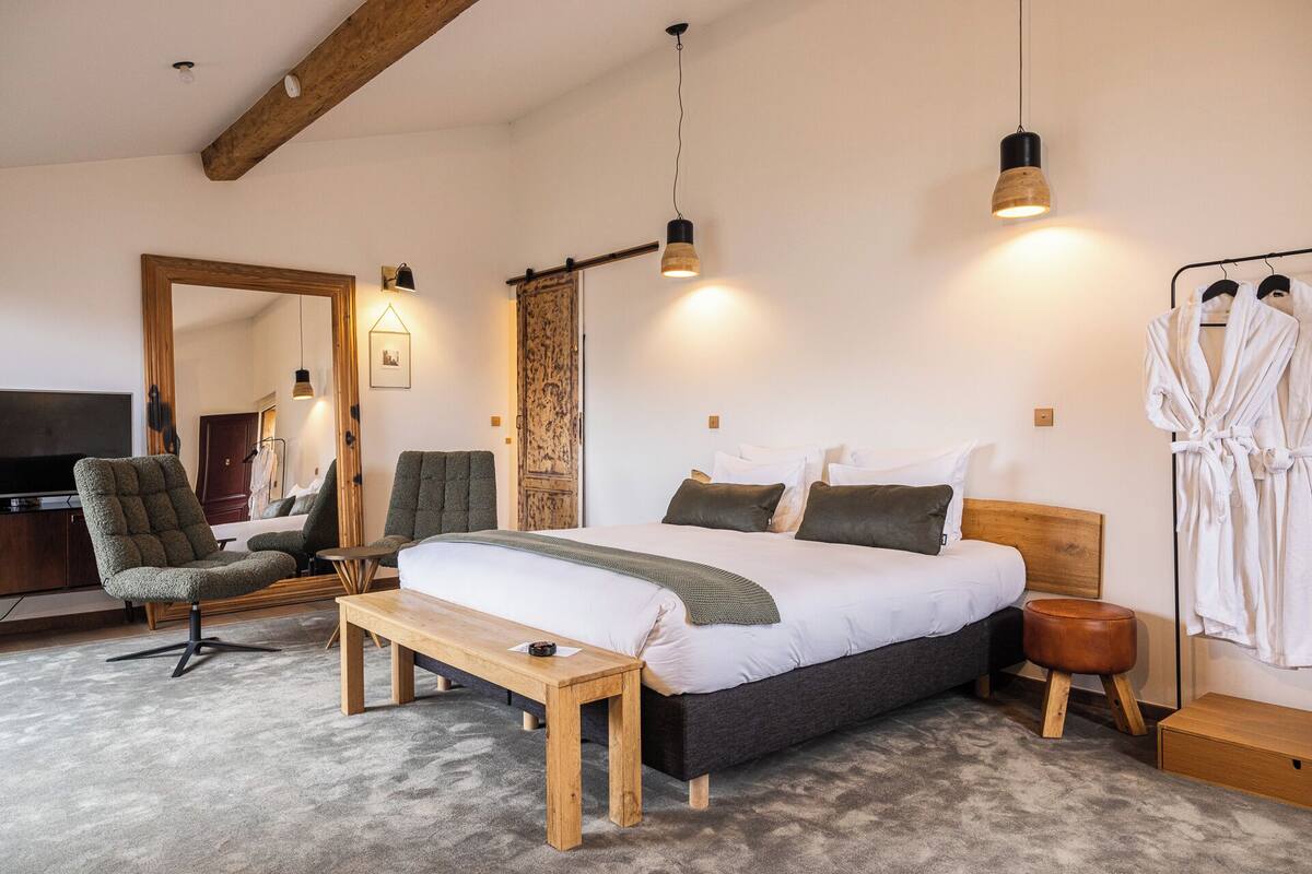 A hotel room with a large bed, a wooden bench, a chair, a TV, and a mirror.