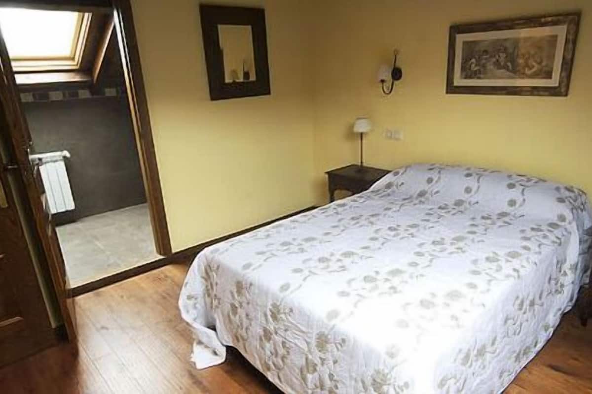 Ein Schlafzimmer mit einem Bett, einem Nachttisch, einer Wandlampe, einem gerahmten Bild und einer Tür zu einem anderen Raum.