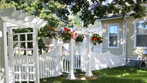 Una pérgola de madera blanca con cestas de flores colgantes, una cerca de piquetes blancos y un césped cuidado frente a una casa.