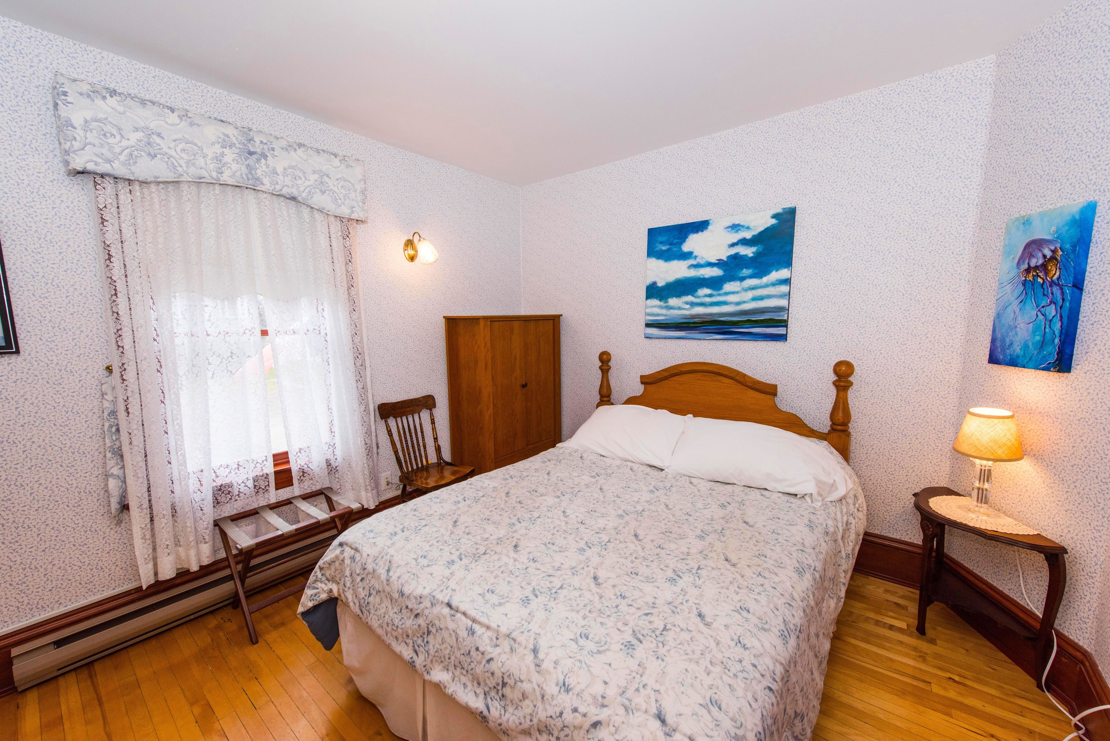 A bedroom with a wooden bed, a nightstand with a lamp, a window with curtains, and a painting on the wall.