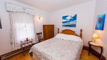 A bedroom with a wooden bed, a nightstand with a lamp, a window with curtains, and a painting on the wall.