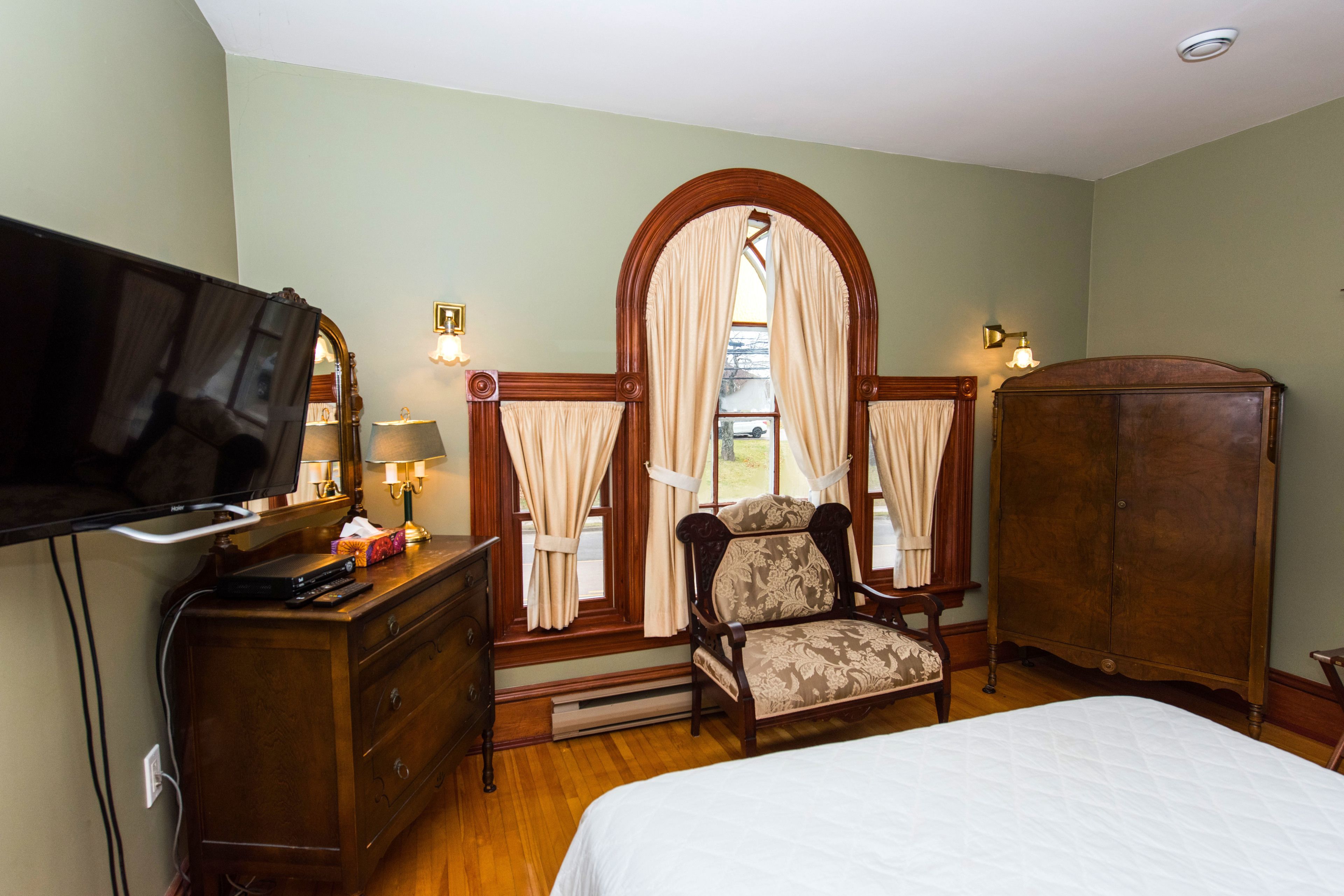A hotel room with a bed, a dresser, a television, a chair, a wardrobe, and a mirror with curtains.