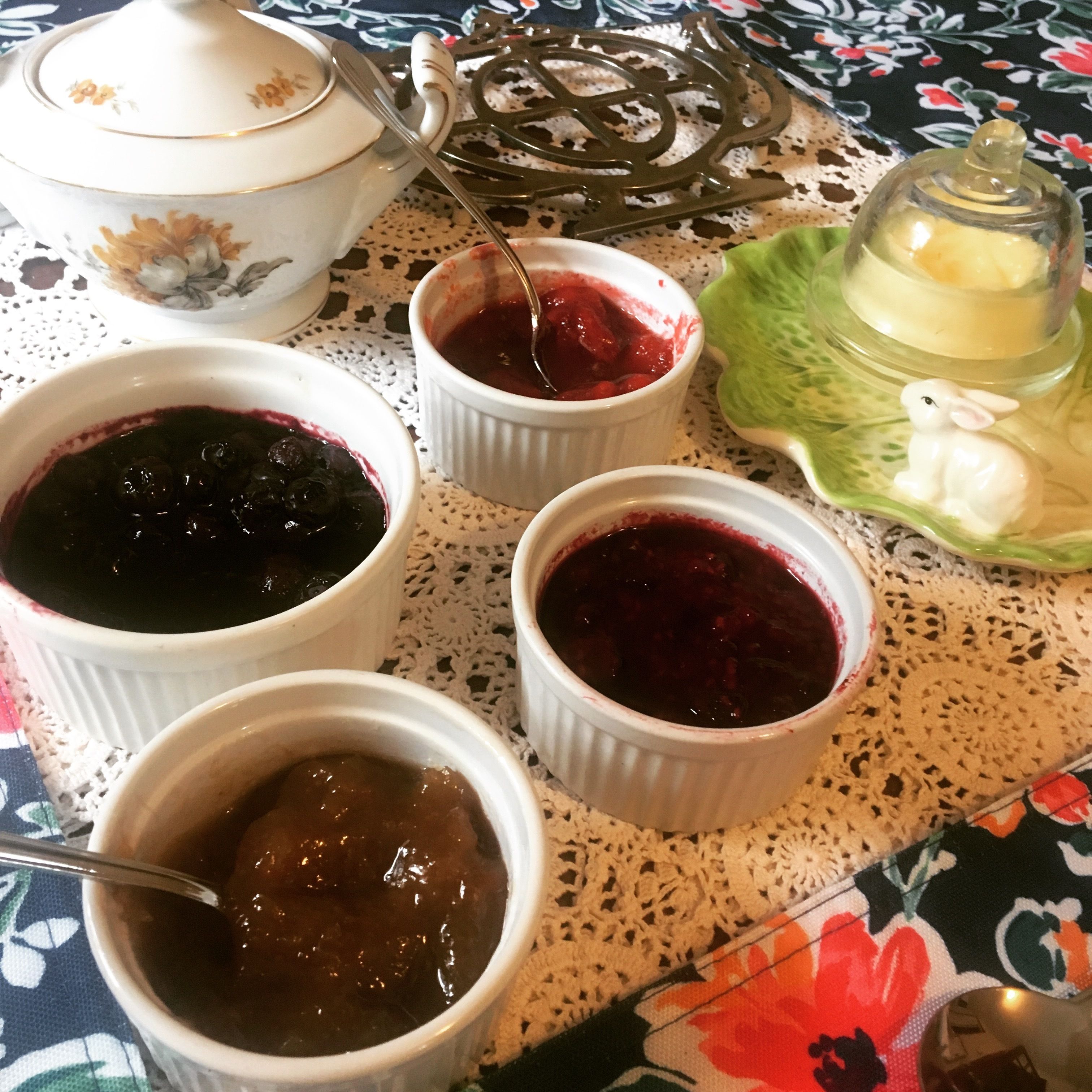 Een tafel met vier witte schaaltjes gevuld met verschillende soorten jam, een groene keramieken schotel met een konijntje en een witte theepot met bloemmotieven.