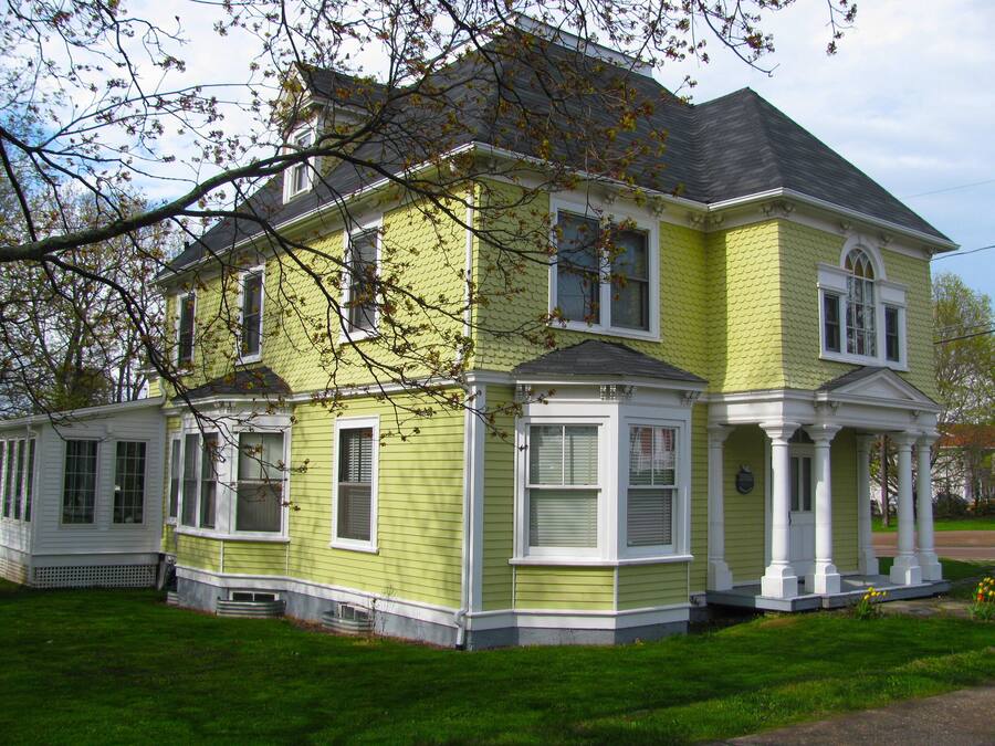 A two-story house with a green exterior and white trim, featuring a porch with columns and a small garden area.