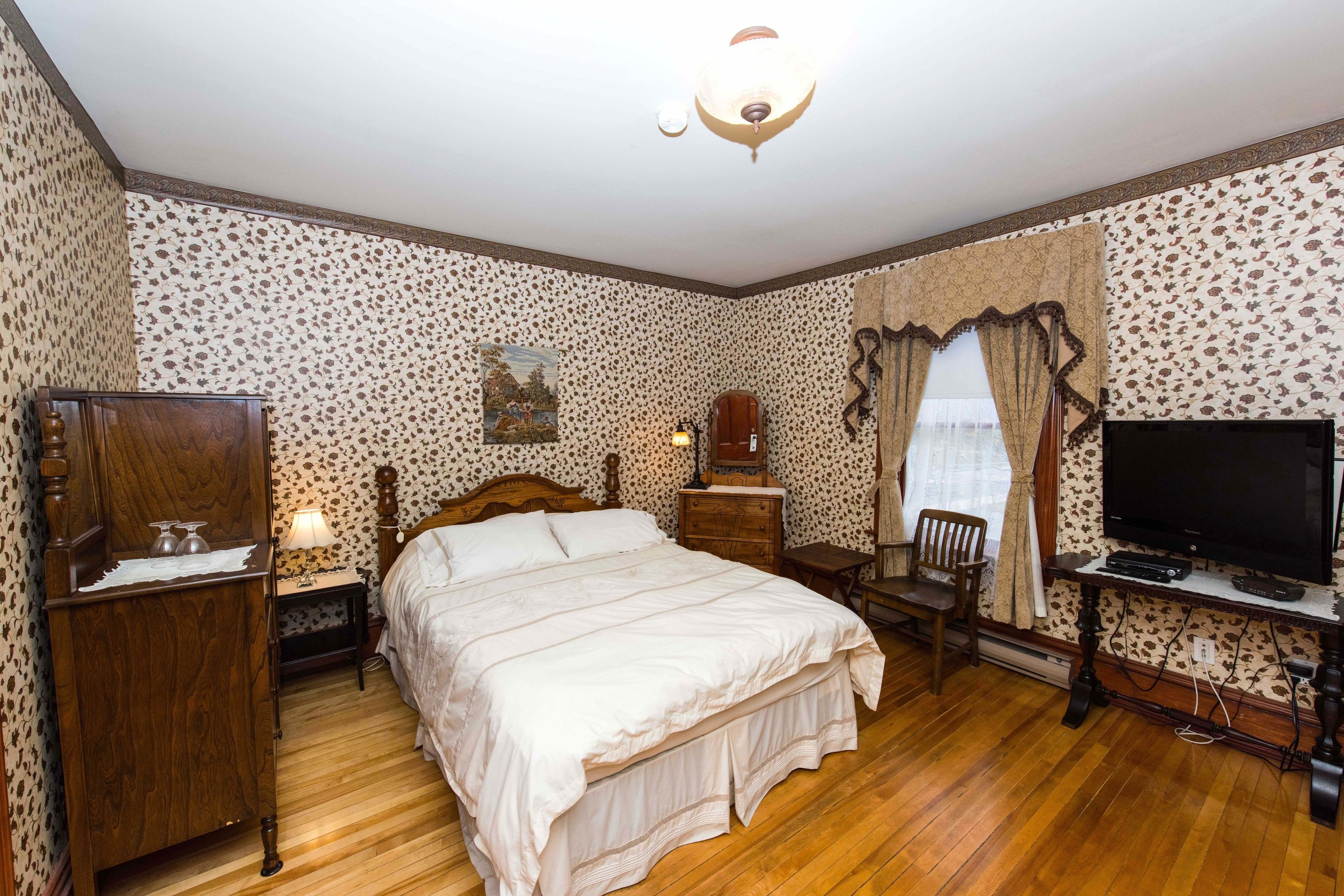 A bedroom with a wooden bed, a nightstand, a dresser, a television, and a window with curtains.