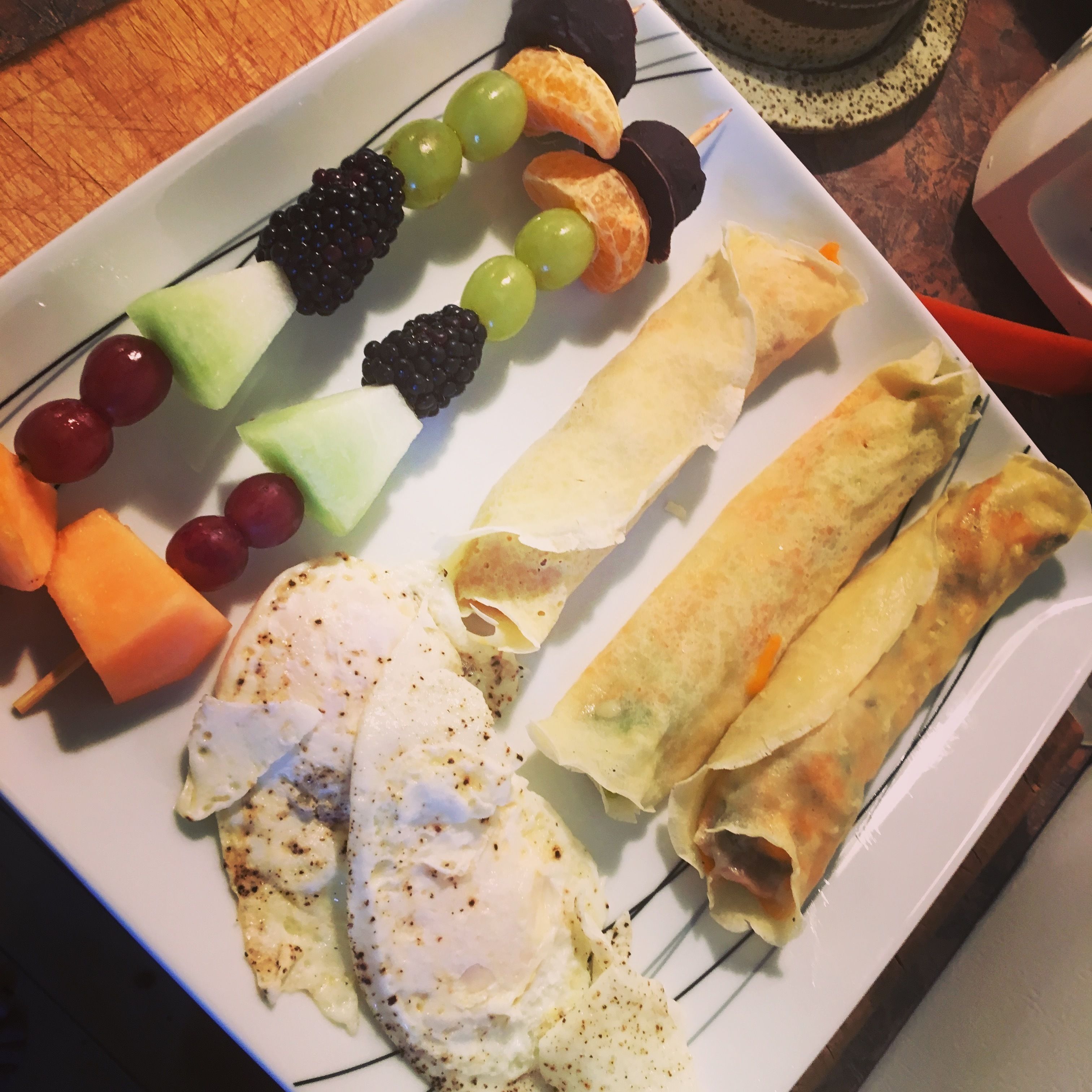 Une assiette contenant divers aliments, notamment des œufs, des brochettes de fruits et des petits pains.