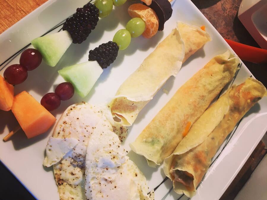 Un plato con diferentes alimentos, como huevos, brochetas de fruta y panecillos.