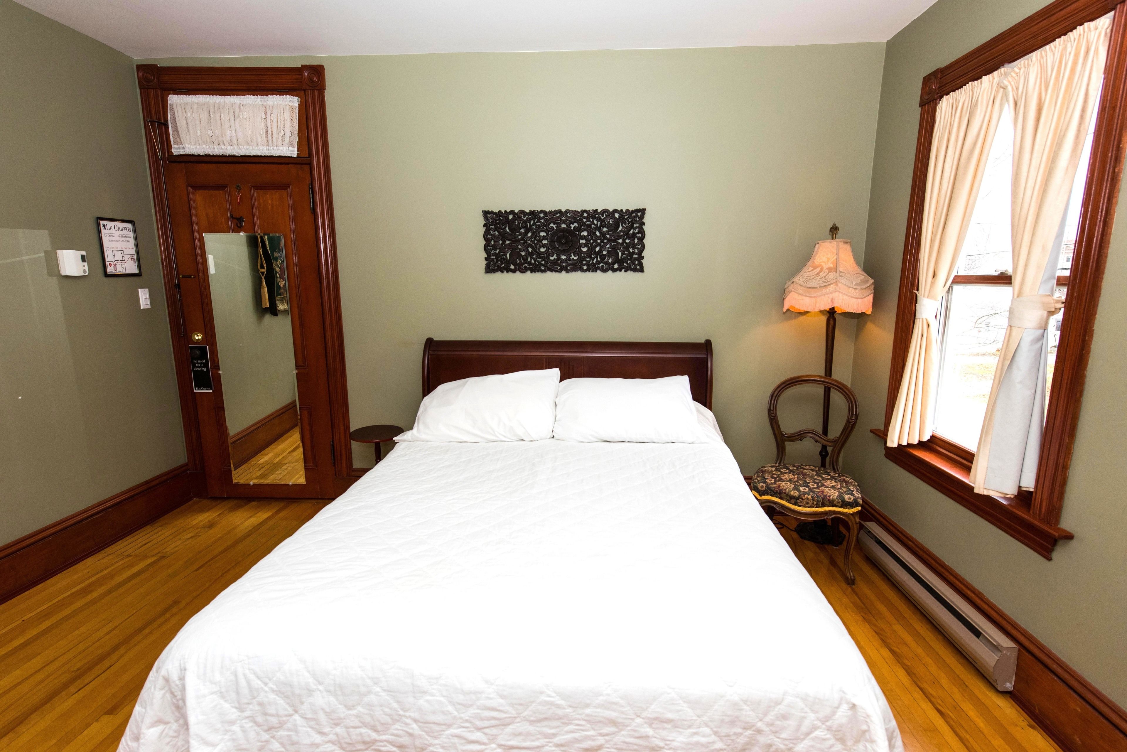 A neatly made bed with white linens, a wooden headboard, a wall-mounted decorative piece, a chair with a cushion, and a window with curtains.