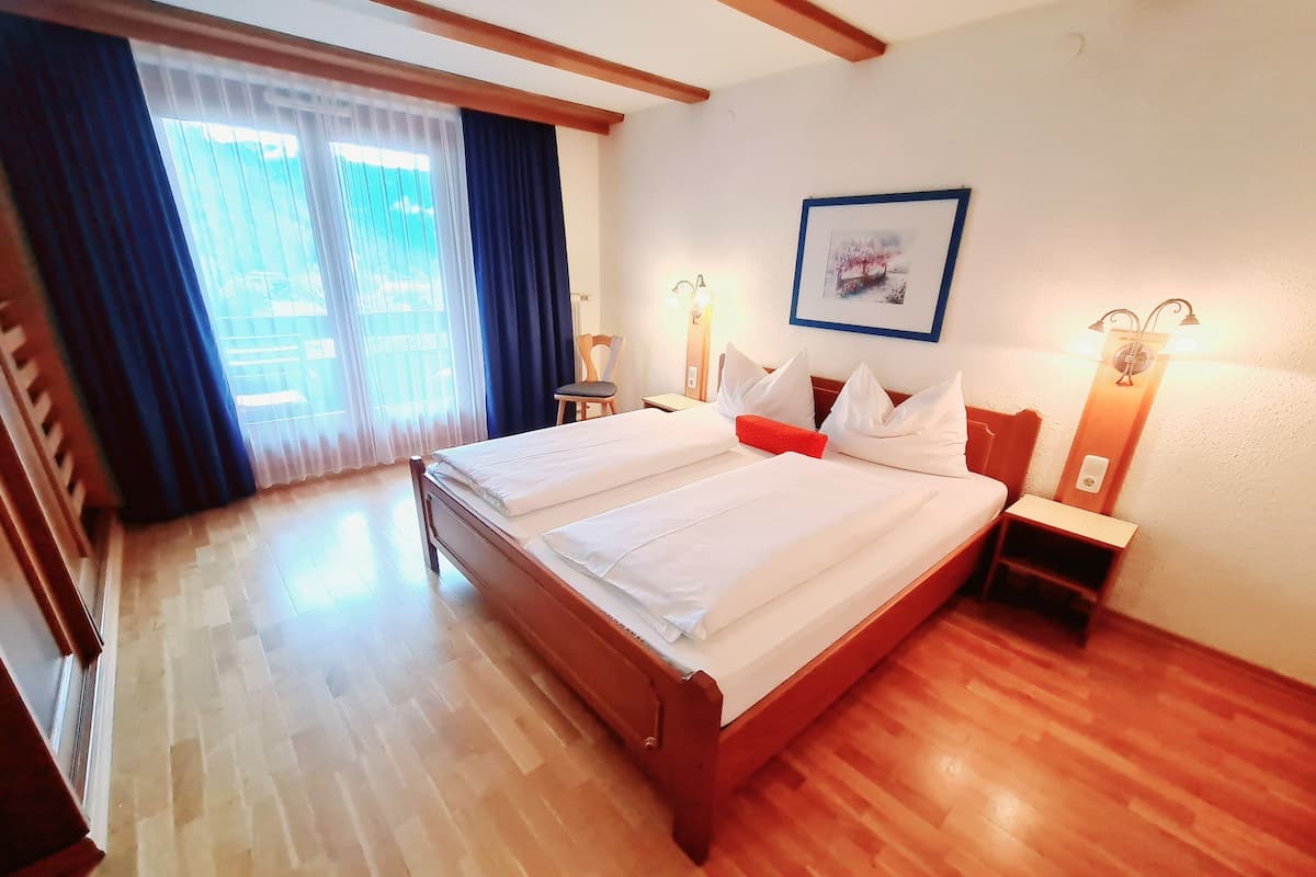 A hotel room with a wooden bed, a nightstand, a chair, a window with curtains, and a wall-mounted picture.