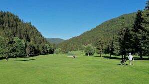 Un campo de golf bajo un cielo despejado, rodeado de bosques densos y colinas.