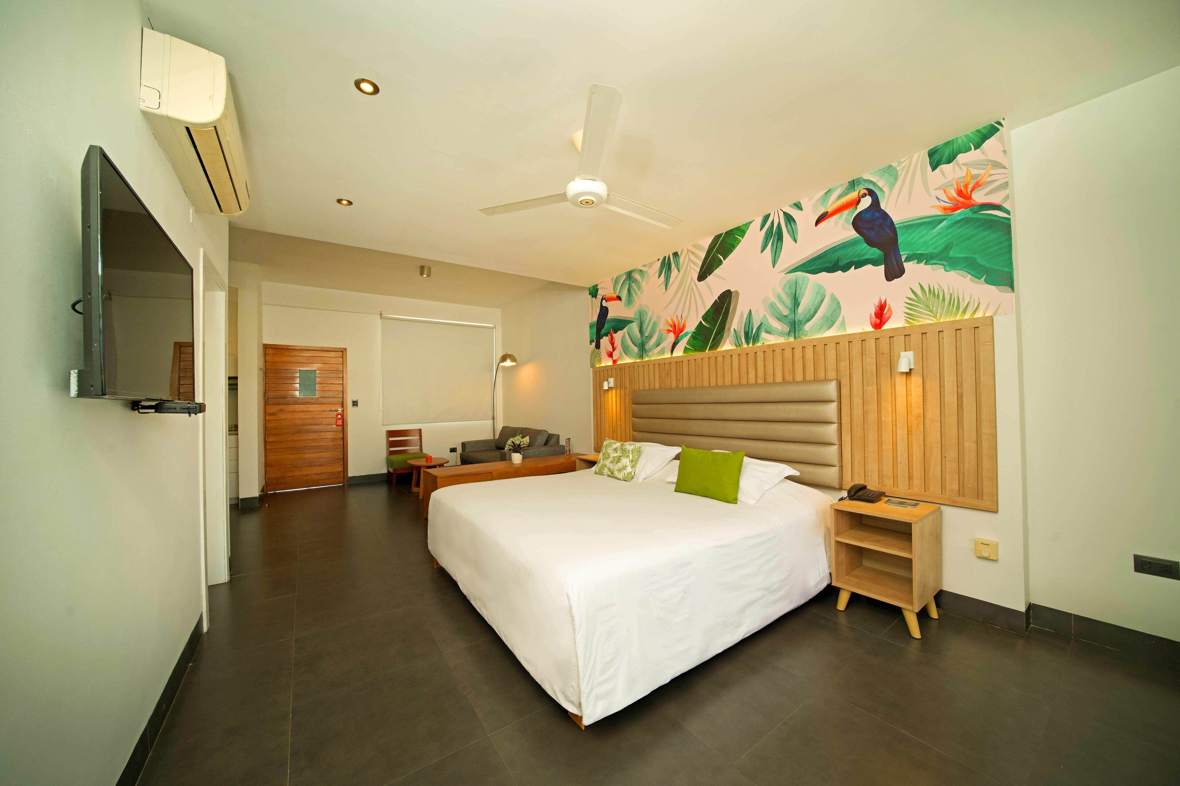 A hotel room with a large bed, a wooden headboard, a wall-mounted TV, and a tropical-themed mural.