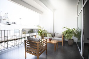 Un balcón moderno con sofá, una silla de madera y una mesa de centro, con plantas en macetas y vistas a la ciudad.