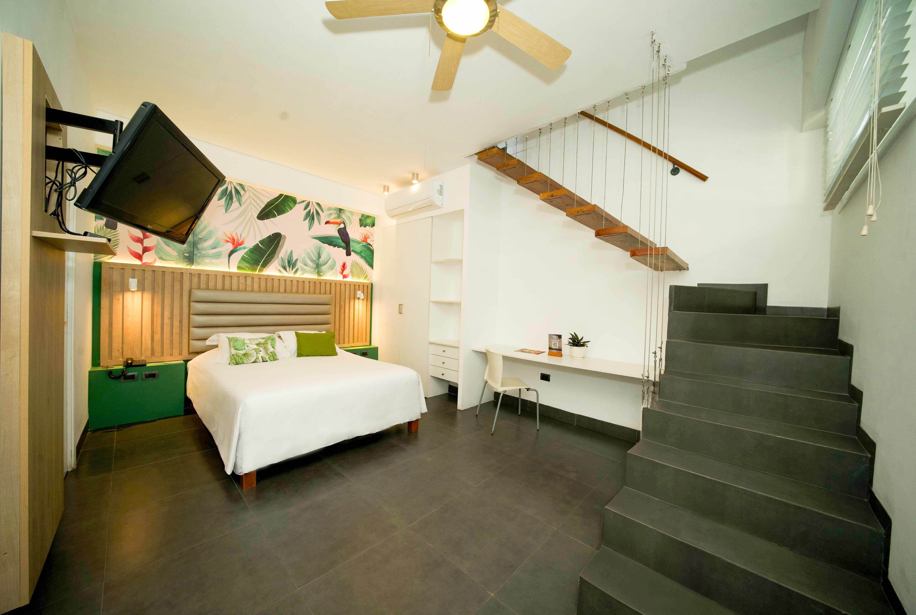 A bedroom with a bed, a desk, a staircase, and a wall-mounted TV.