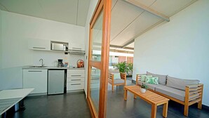 Cozinha moderna com armários brancos, micro-ondas embutido e máquina de café. Há uma porta deslizante com moldura de madeira que dá para uma sala de estar com sofá, mesa de centro e planta.