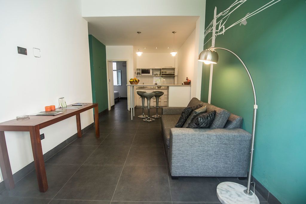 Una sala moderna con un sofá gris, una mesa de centro de madera y una lámpara de pie. Se ve una cocina al fondo con un taburete de bar y un frutero.