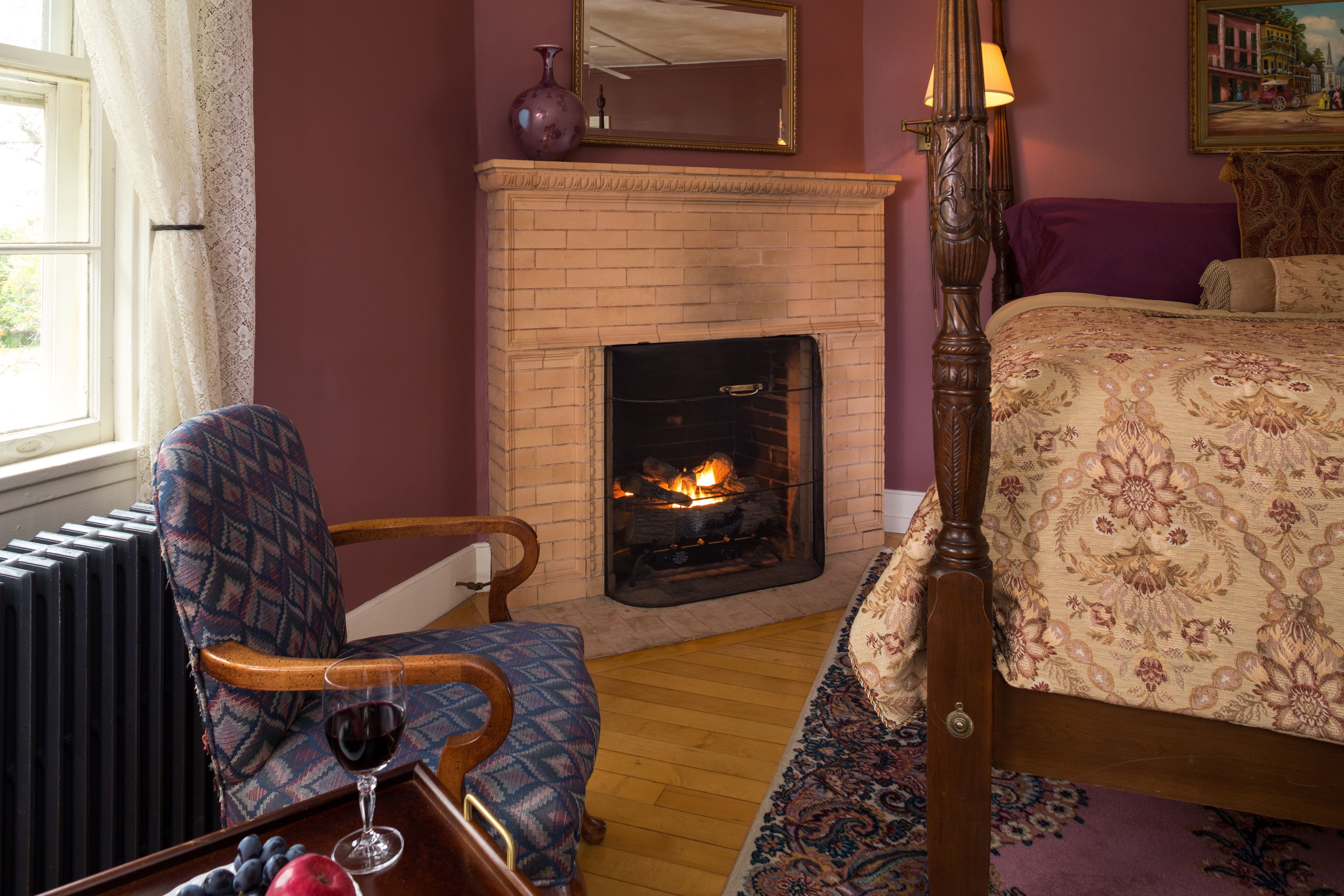 A bedroom with a brick fireplace, a bed with floral bedding, a chair with a patterned cushion, and a table with a glass of red wine and fruit.