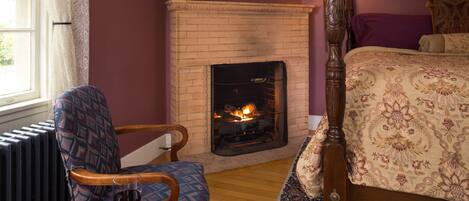 A bedroom with a brick fireplace, a bed with floral bedding, a chair with a patterned cushion, and a table with a glass of red wine and fruit.