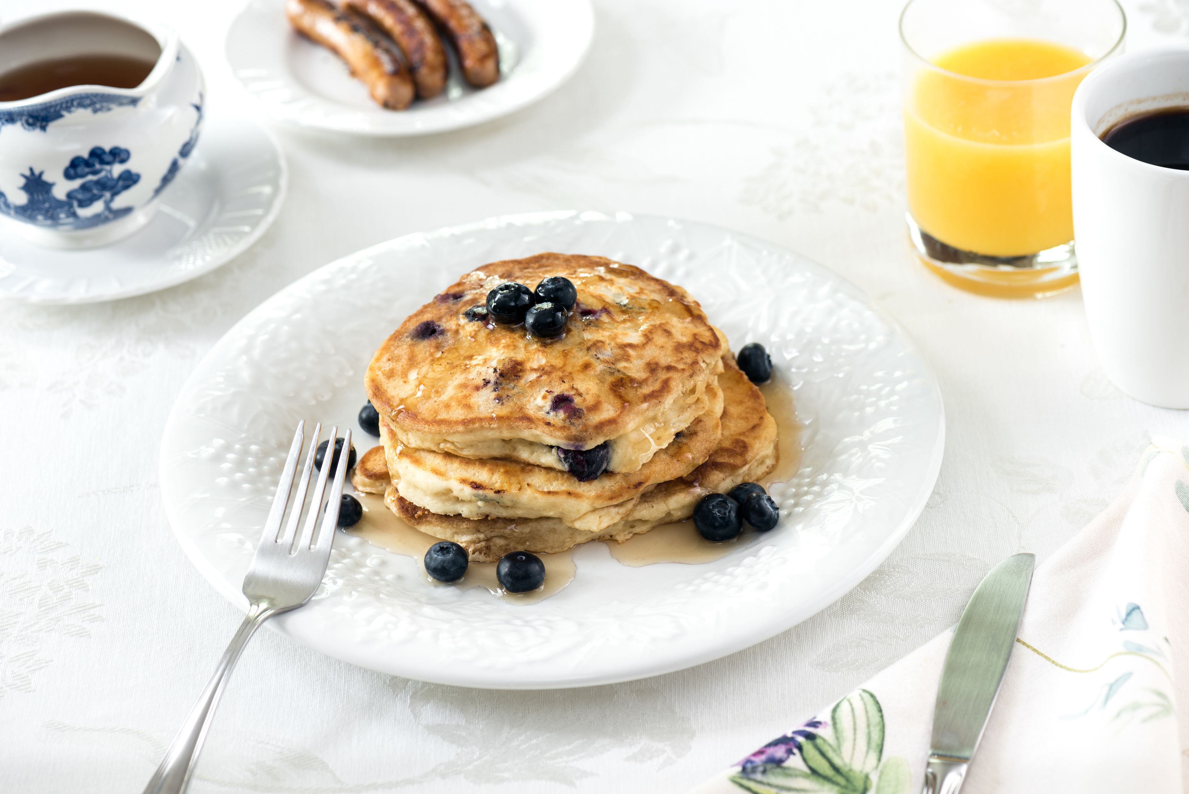 A breakfast plate with pancakes, blueberries, and syrup, a cup of coffee, and a glass of orange juice on a table.