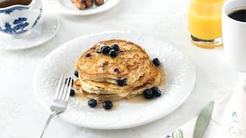 A breakfast plate with pancakes, blueberries, and syrup, a cup of coffee, and a glass of orange juice on a table.