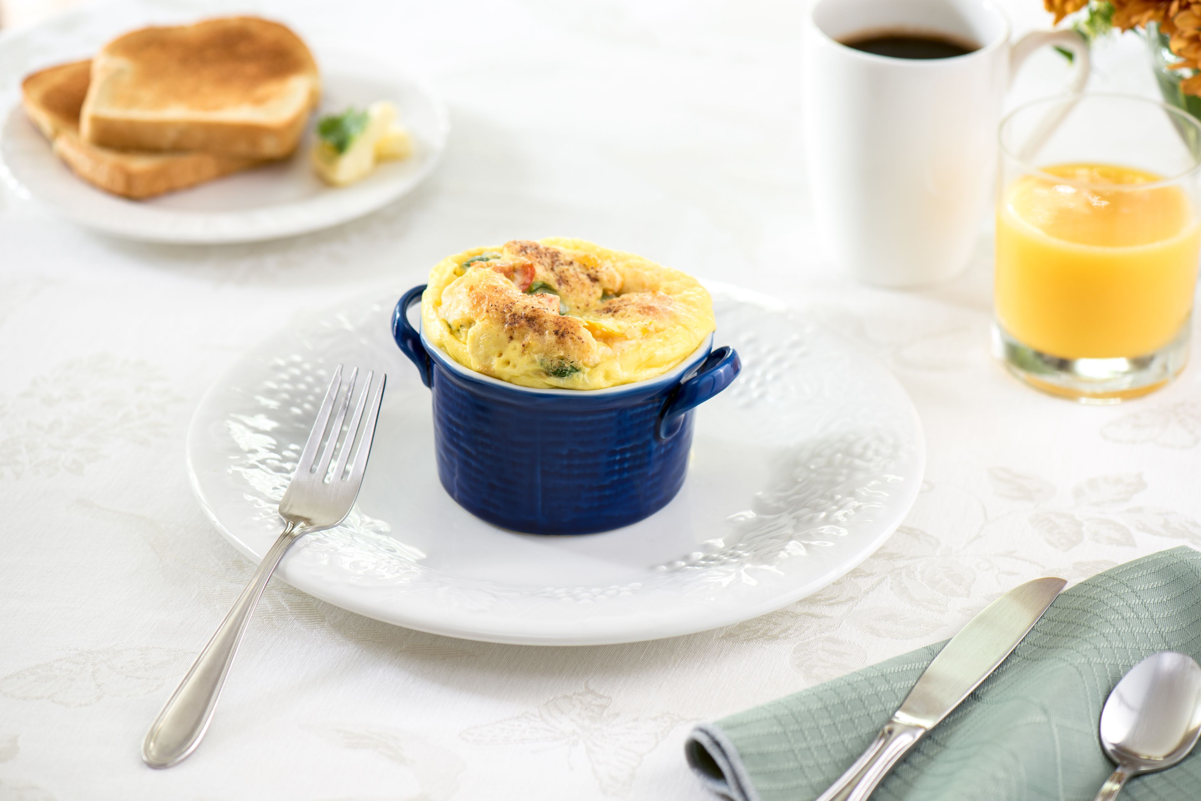 A breakfast setting with a blue ramekin containing a dish, a glass of orange juice, and toast on a white plate.