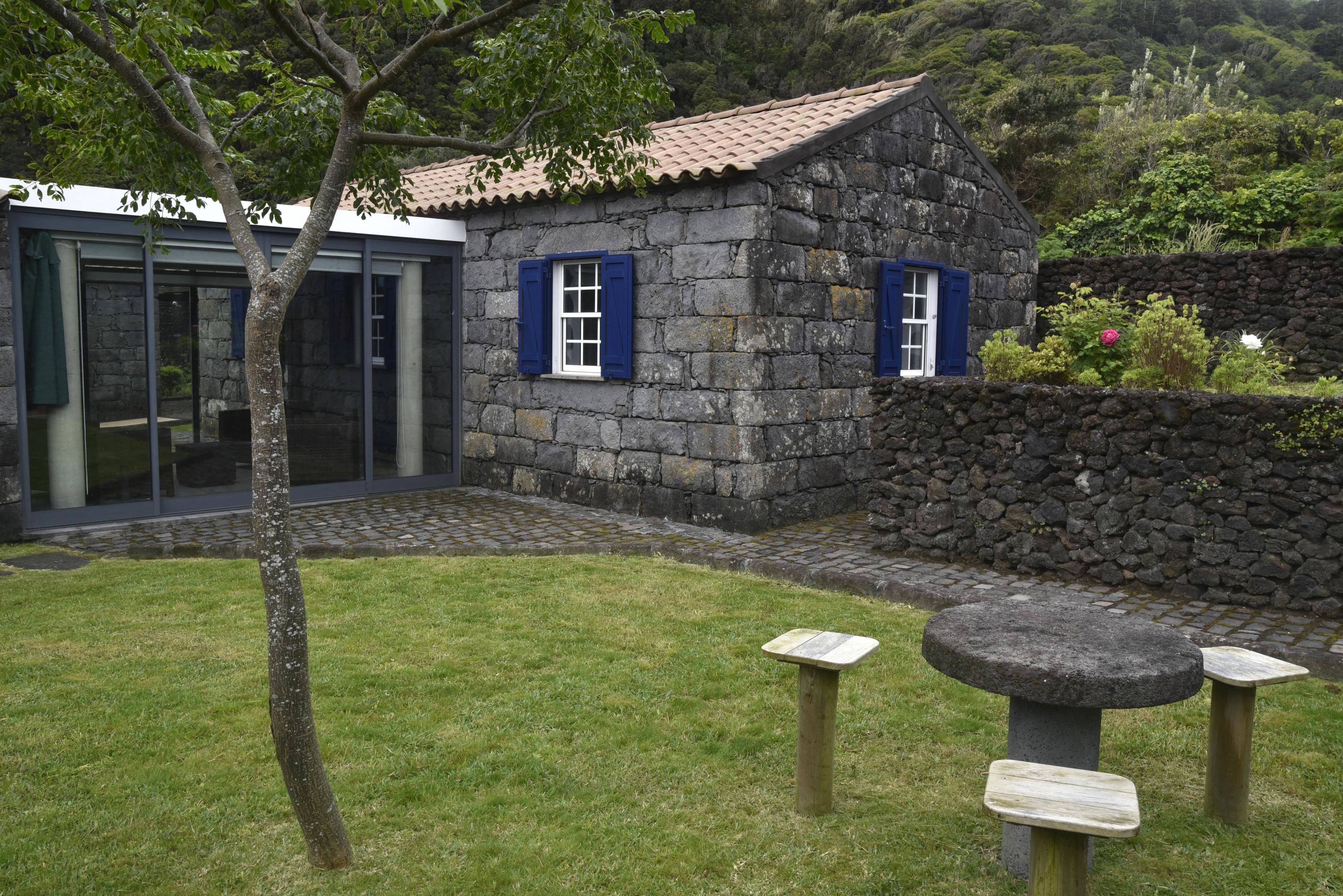 Una casa de piedra con contraventanas azules, un muro de piedra y un patio con césped y un banco de piedra.