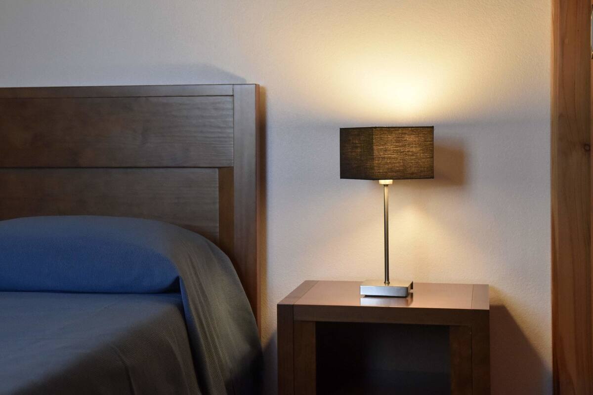 Un lit bien fait, recouvert d’une couverture bleue, avec une tête de lit en bois et une petite table de chevet surmontée d’une lampe.