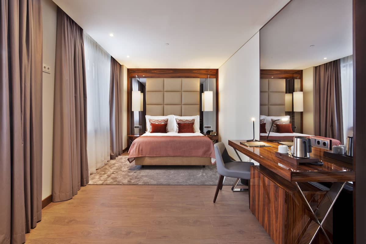A modern hotel room with a large bed, a desk with a chair, a mirror, and a small table with a kettle.