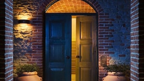 Un arco de ladrillo que conduce a una puerta de madera, con un muro de piedra a un lado y una planta en maceta al otro.