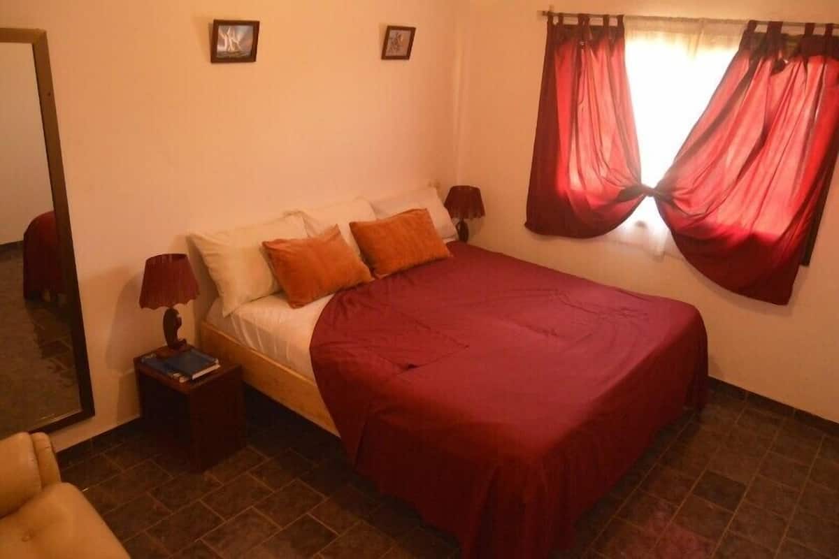 A bedroom with a bed, pillows, a nightstand, a lamp, a chair, and a window with curtains.
