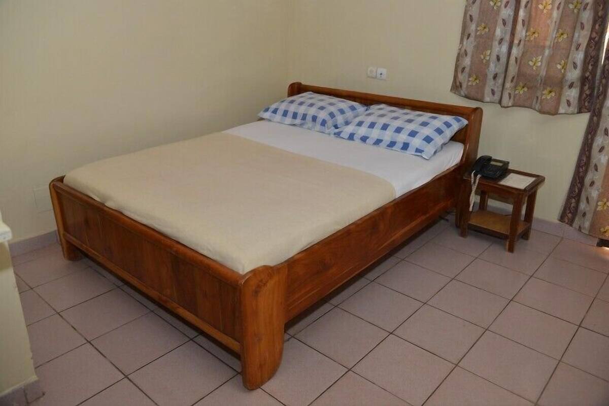 A wooden bed with a white mattress and blue checkered pillows, a bedside table with a phone, and a curtain with a floral pattern.