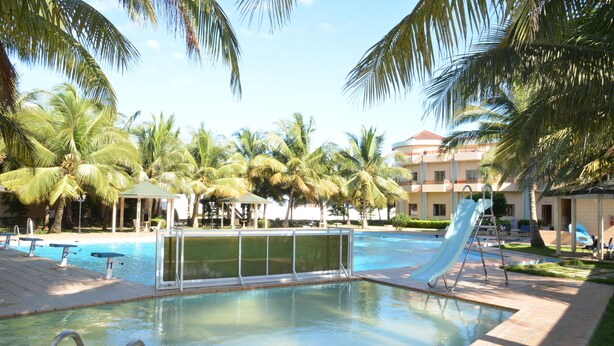 Une piscine avec un toboggan, entourée de palmiers et bordée d’un bâtiment au toit rouge.