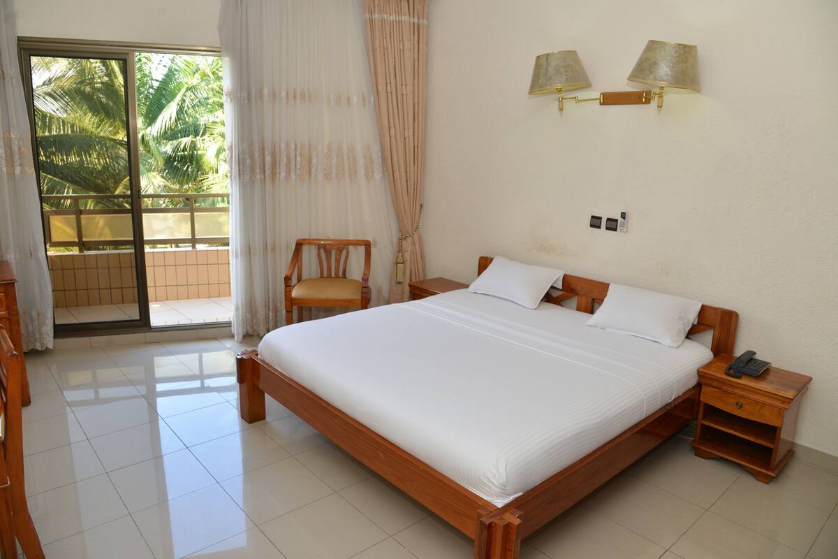 Quarto com cama de madeira, mesa de cabeceira, cadeira, janela com cortinas e vista para palmeiras.
