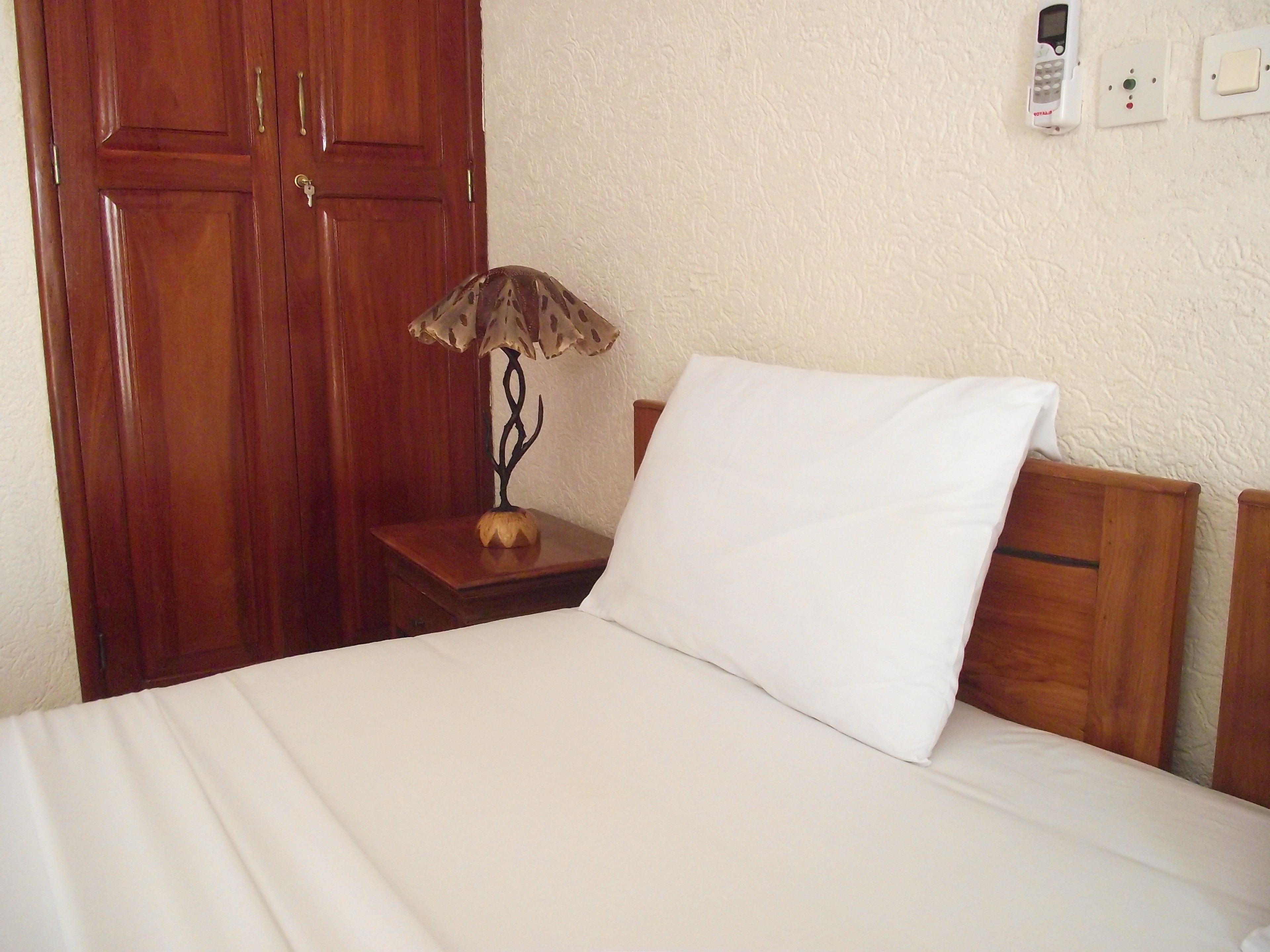 A hotel room with a wooden wardrobe, a bedside table with a lamp, and a white bed with a pillow.
