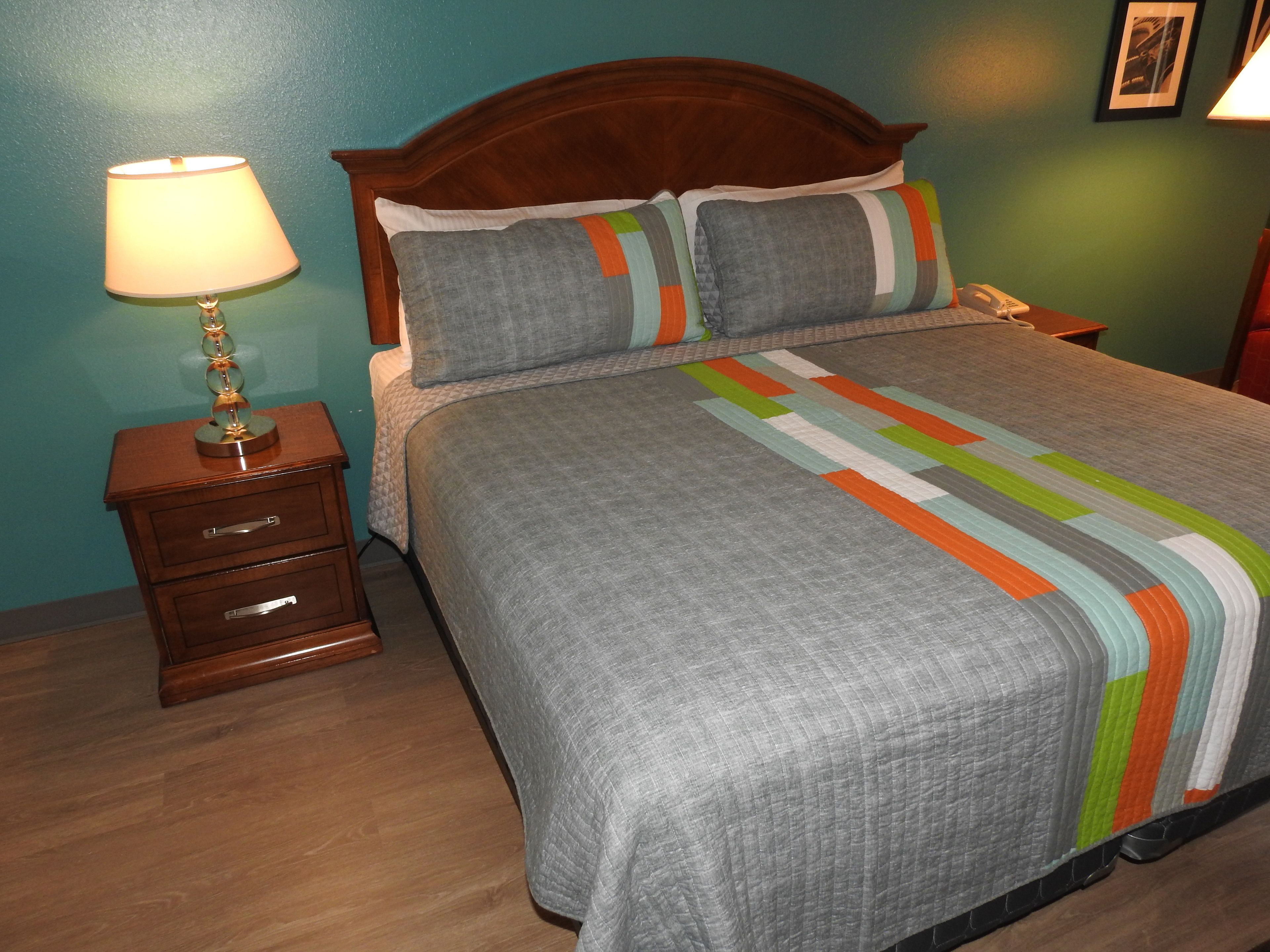 Una cama bien hecha con colcha gris y rayas coloridas, una mesita de noche de madera con una lámpara, y un aparador de madera con cajones.