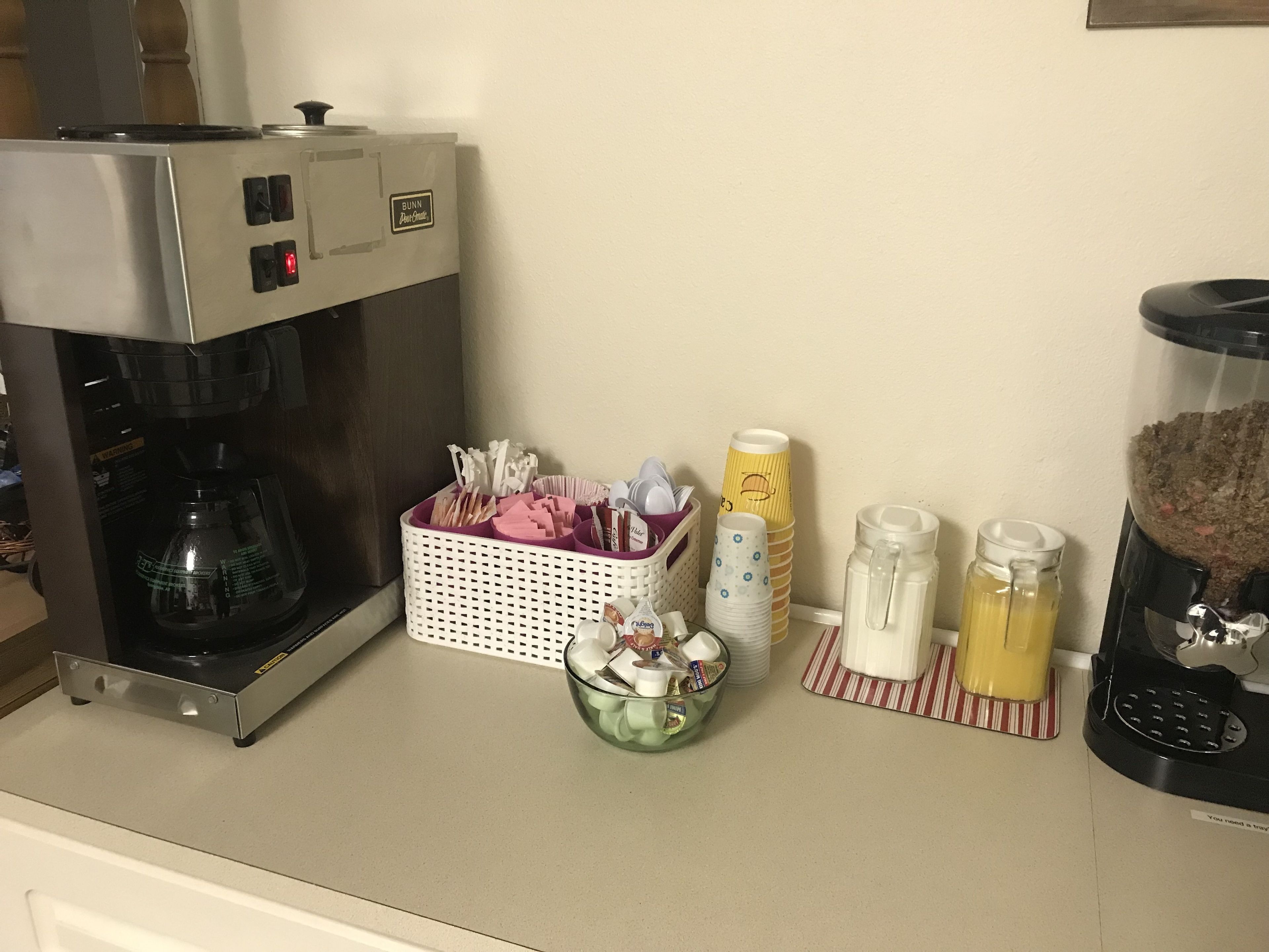 A kitchen area with a coffee maker, a basket of utensils, and a coffee station with cups and a milk jug.