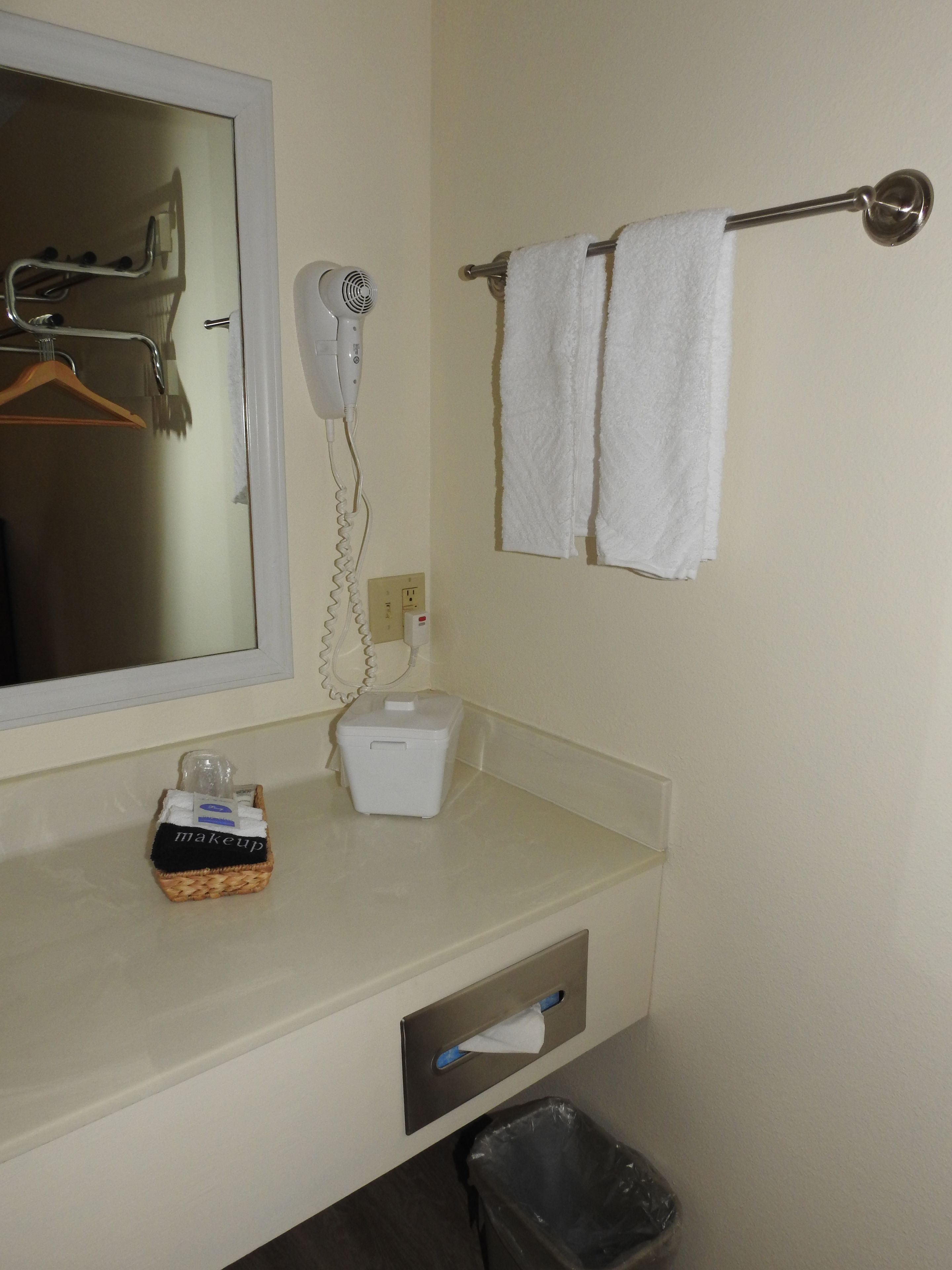 A bathroom with a mirror, a hairdryer, a towel rack with two towels, a basket with toiletries, and a trash bin.