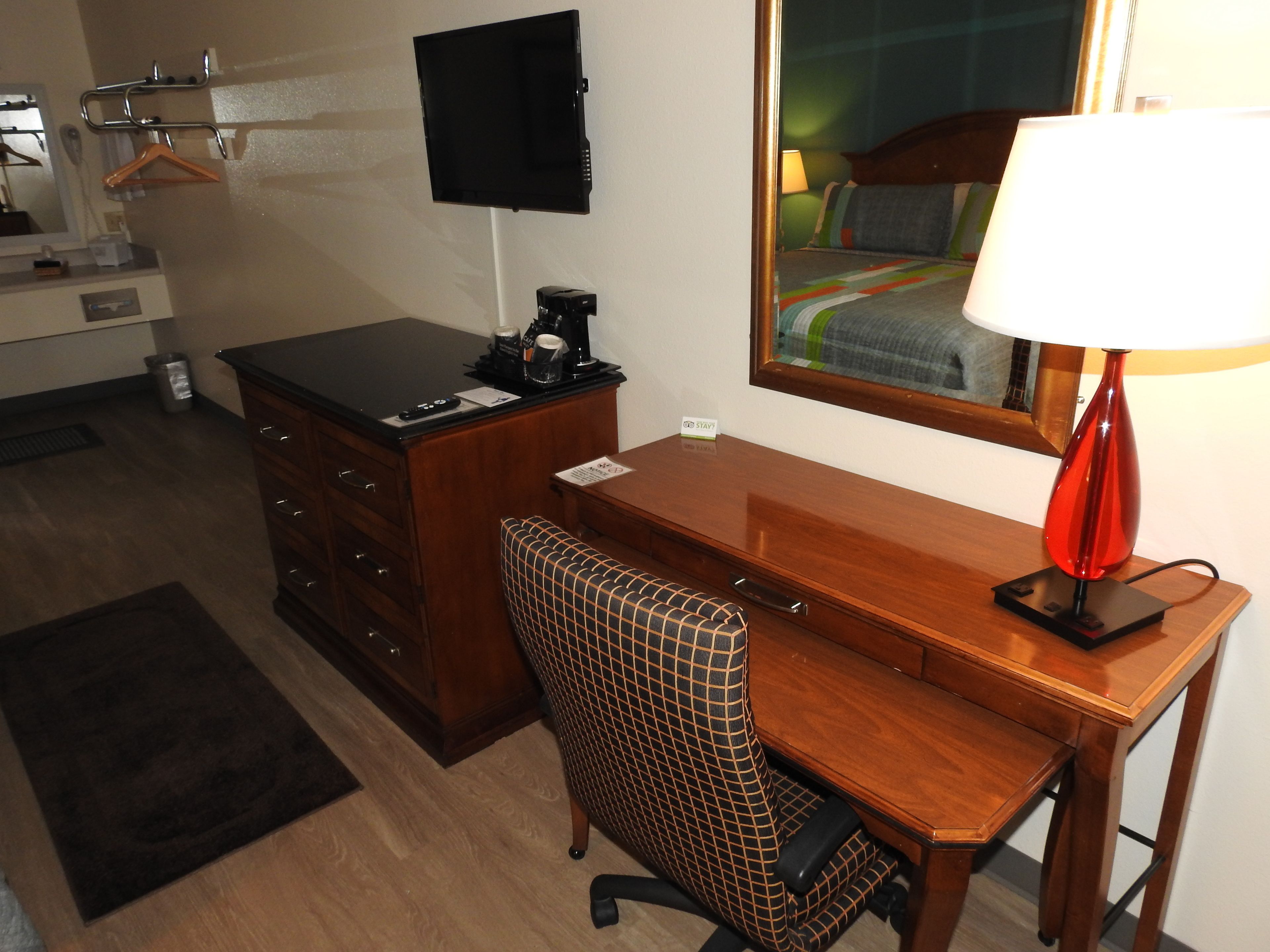 Habitación de hotel con un escritorio de madera, una silla, un televisor montado en la pared, un espejo y una lámpara.