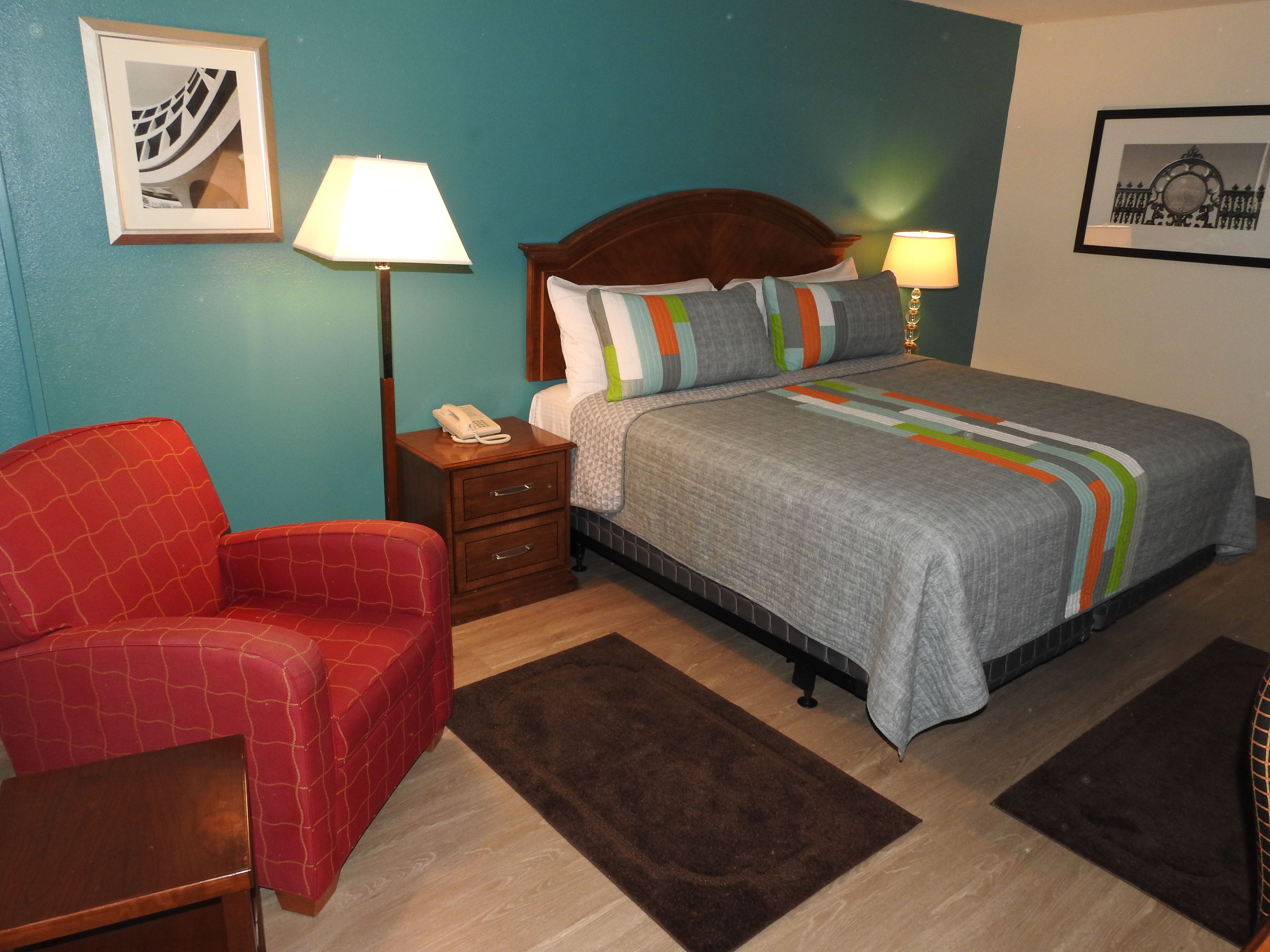 Quarto de hotel com uma cama, uma poltrona vermelha, uma mesinha de cabeceira com abajur e um telefone.