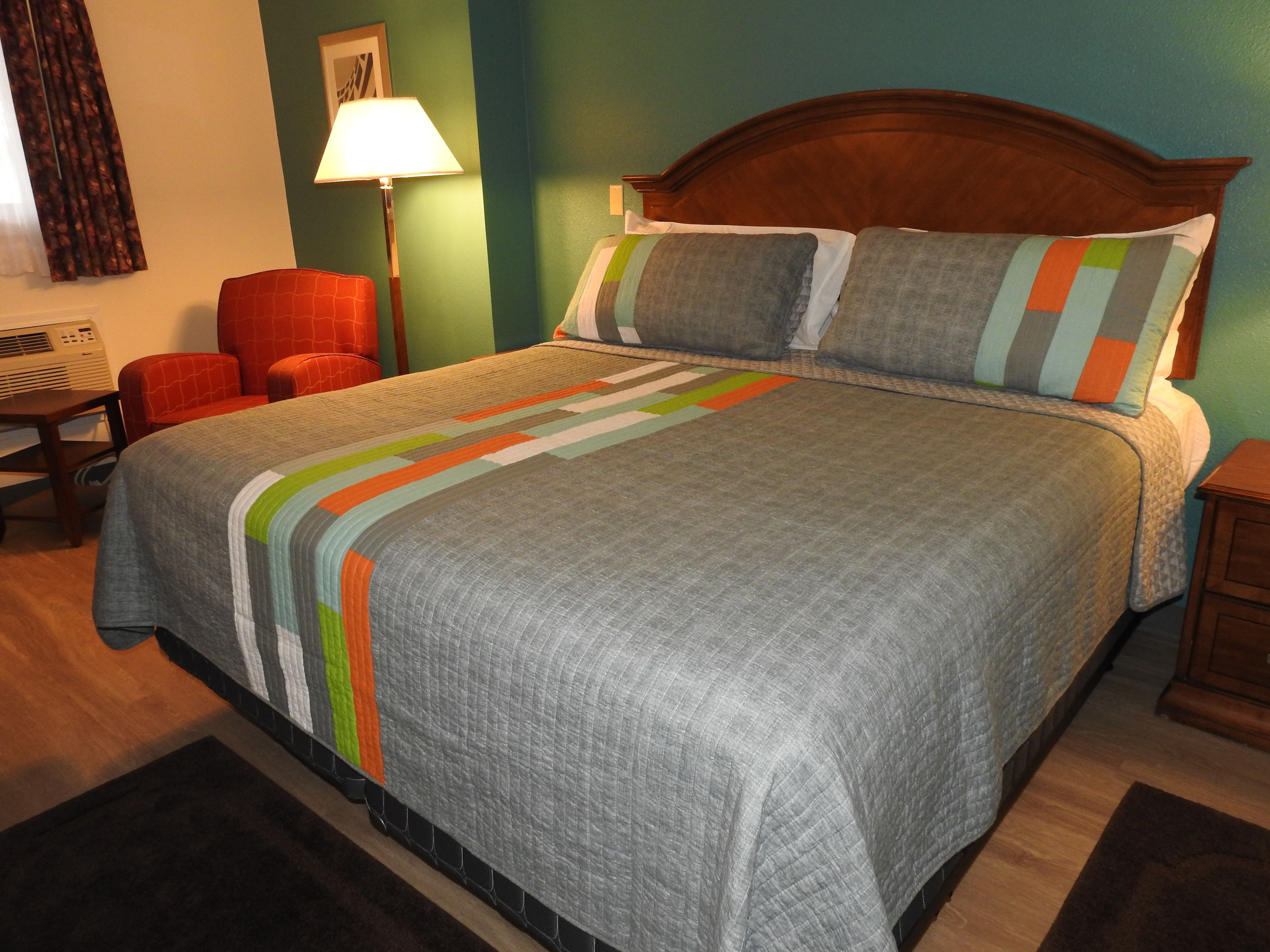 Una cama bien hecha con una colcha a rayas, un cabecero de madera y dos almohadas. Hay una silla roja y una lámpara al lado de la cama.