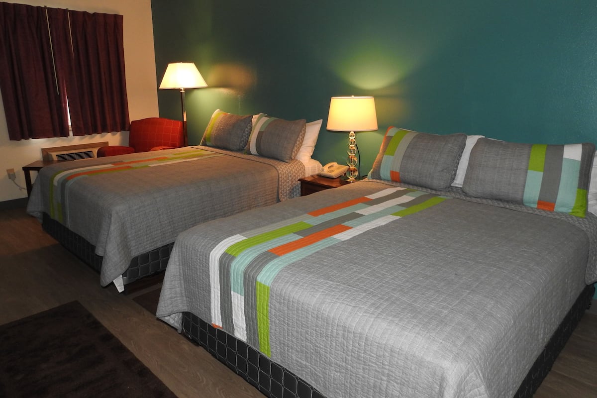 Une chambre d’hôtel avec deux lits, chacun recouvert d’une couette grise, une table de chevet avec une lampe et une chaise rouge.