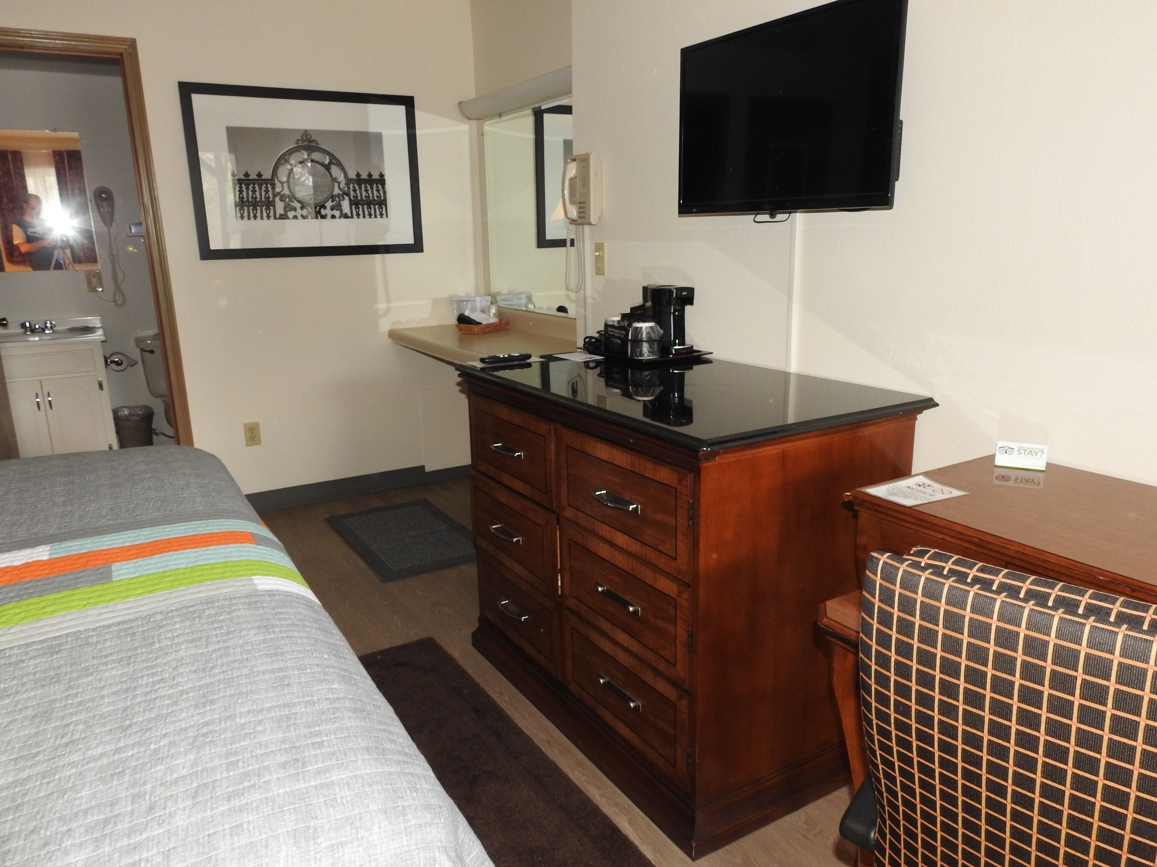 A hotel room with a bed, a wooden dresser, a desk, a chair, a television, a framed picture, a mirror, and a bathroom visible through an open door.