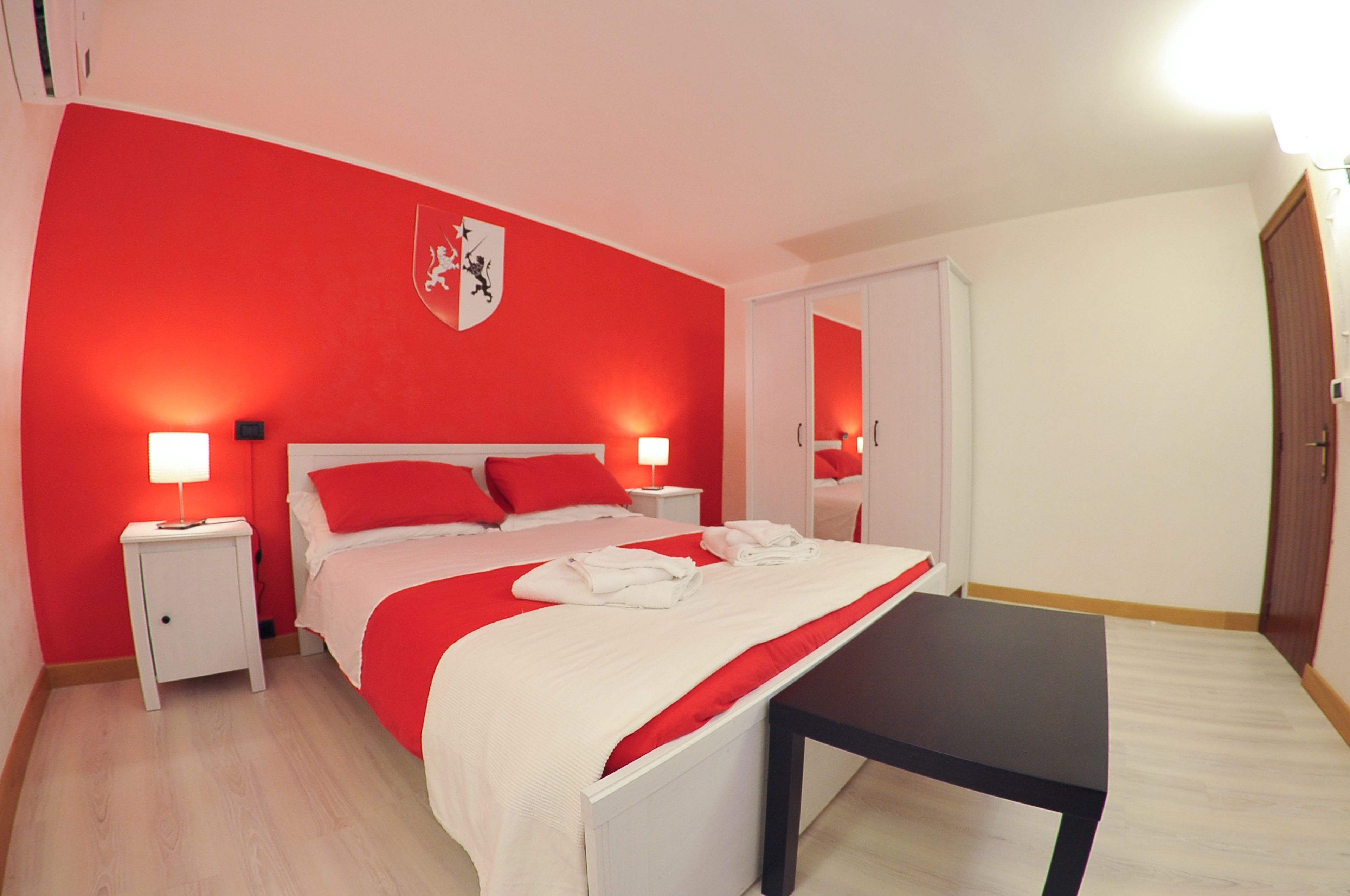 A hotel room with a red and white color scheme, a bed with red bedding, a bedside table with a lamp, and a coat rack.