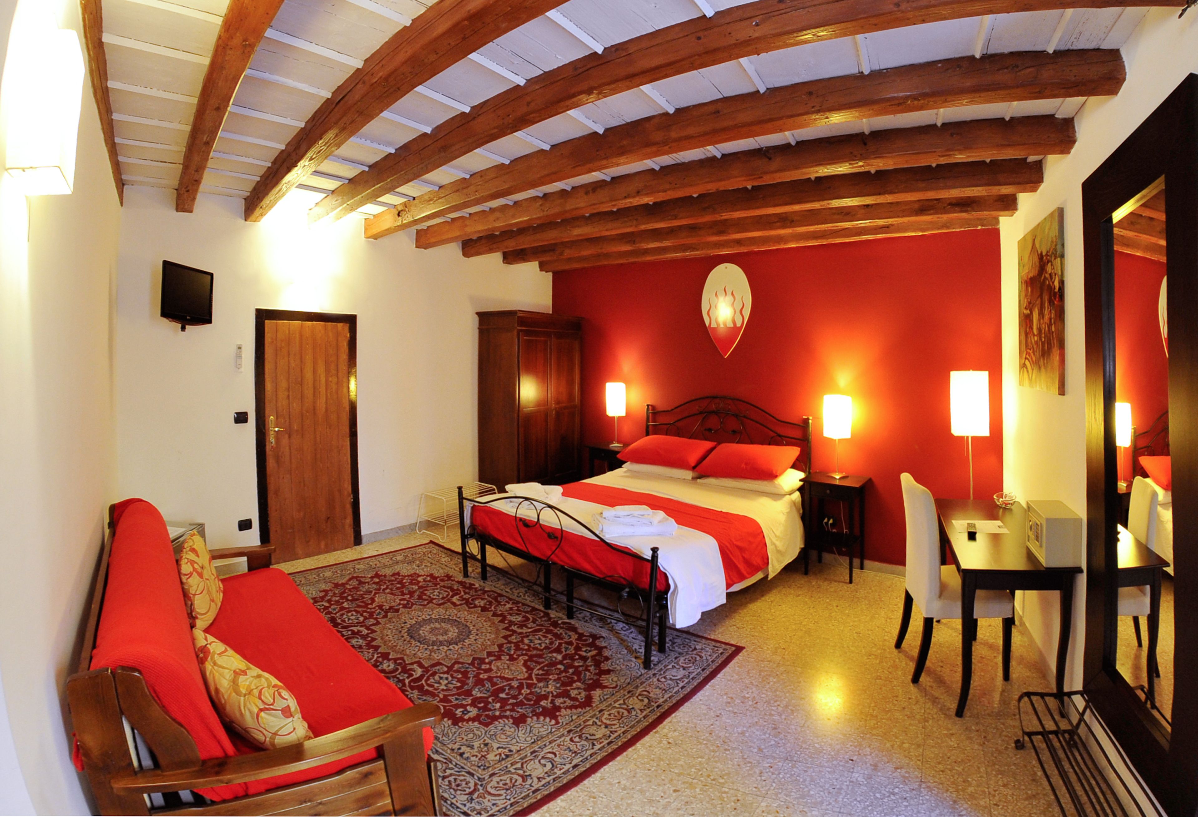 A bedroom with a bed, a desk, a chair, a television, and a red accent wall.