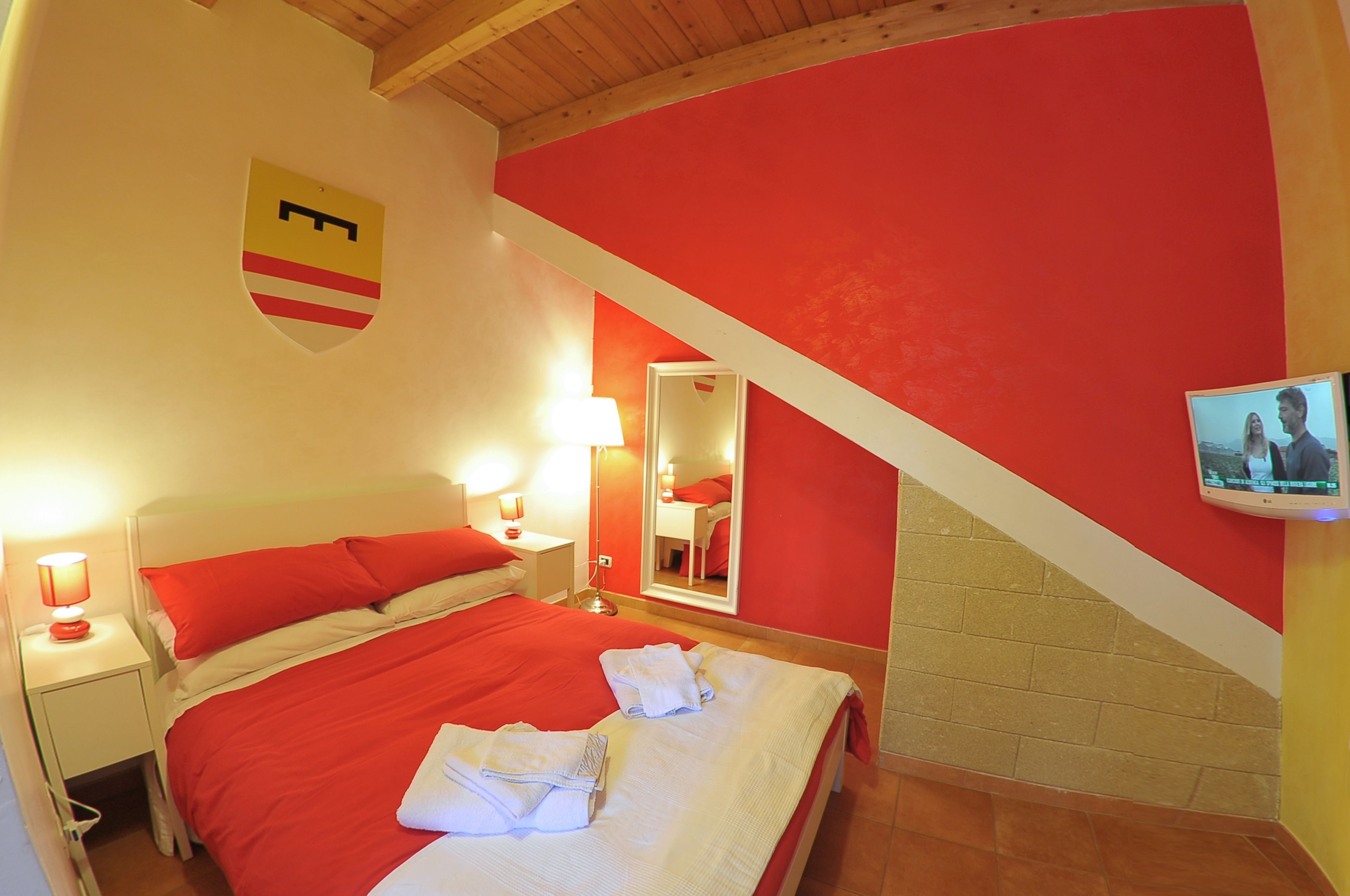 Une chambre d’hôtel aux couleurs rouge et blanc, avec un lit recouvert d’une literie rouge, une table de chevet avec une lampe, un miroir et un téléviseur fixé au mur.
