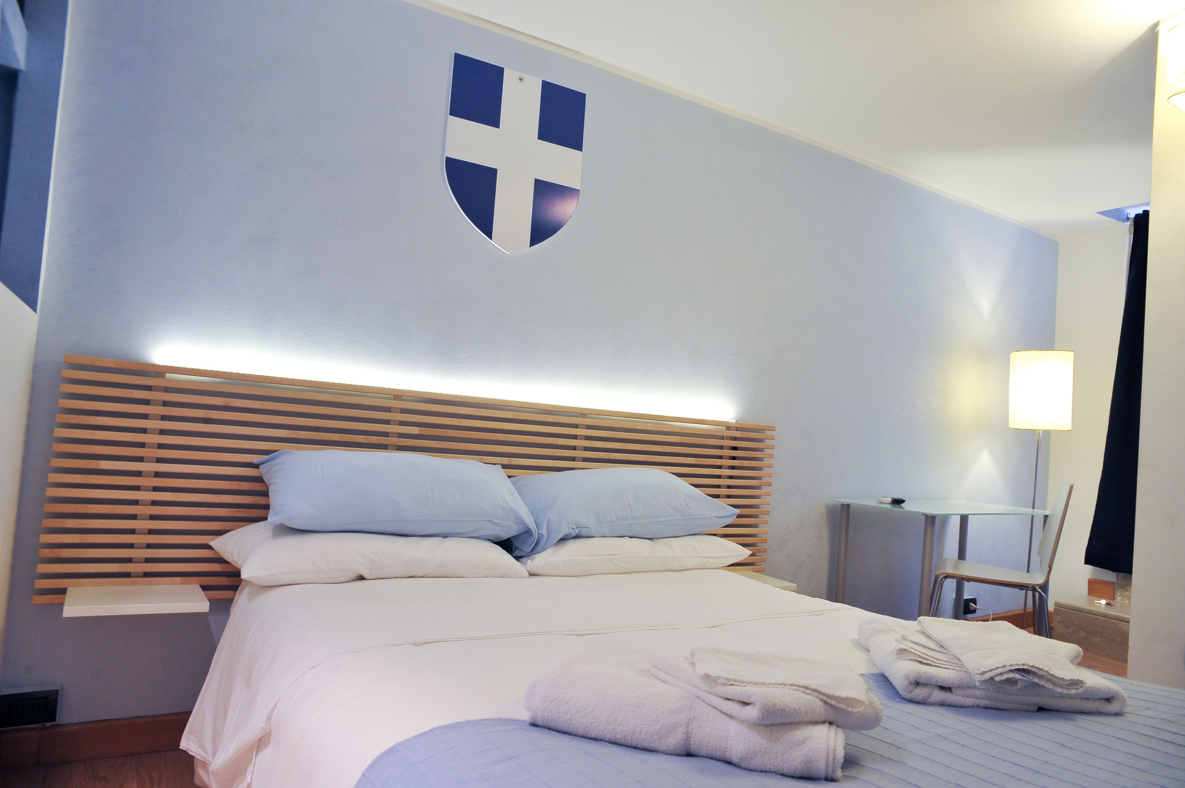 A hotel room with a bed, pillows, a wooden headboard, a desk, a chair, and a wall-mounted coat rack.