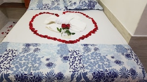 Una cama con un arreglo de pétalos de rosa en forma de corazón, una rosa solitaria, una toalla blanca y una tarjeta sobre el cubrelecho.