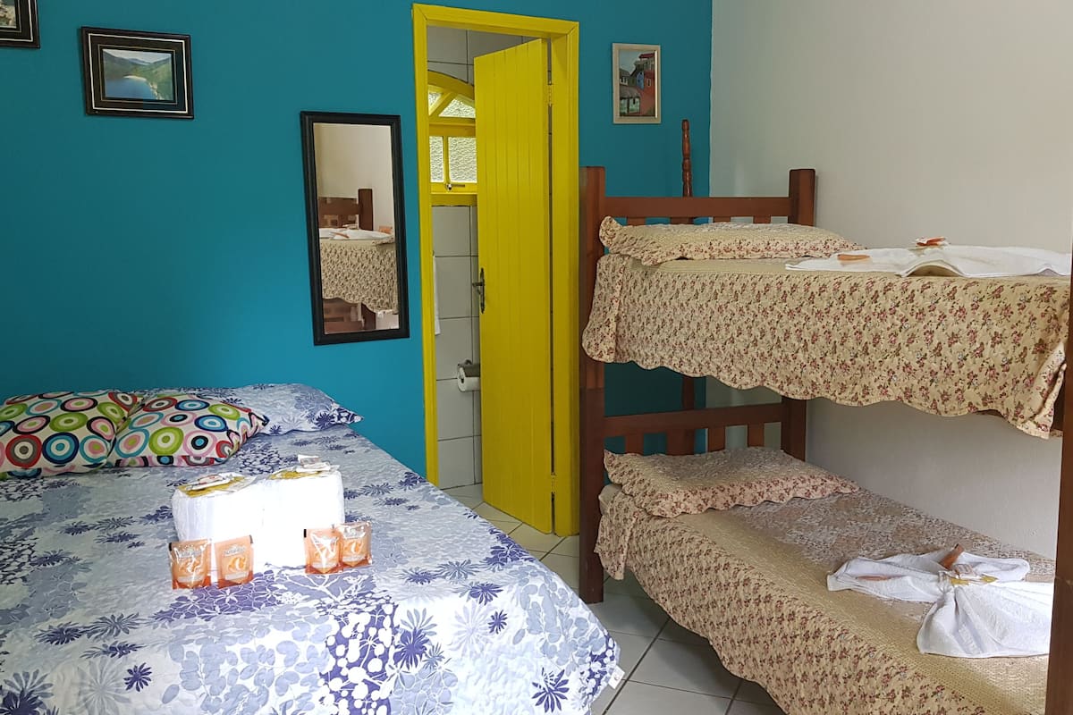 A room with a bunk bed, a single bed with a floral bedspread, a colorful pillow, and a mirror on the wall.