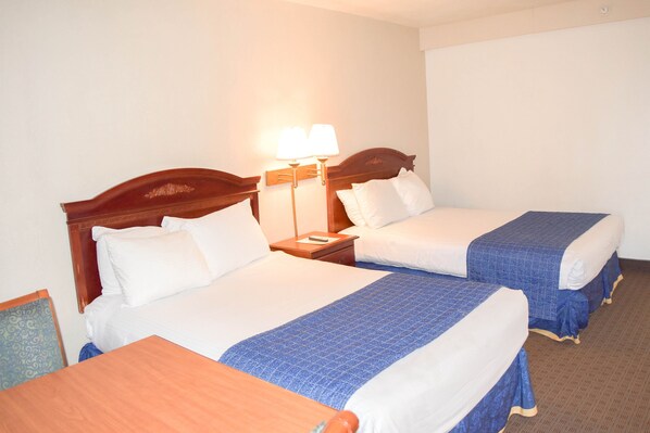 Habitación de hotel con dos camas, un cabecero de madera, una mesita de noche con lámpara y una pequeña mesa.