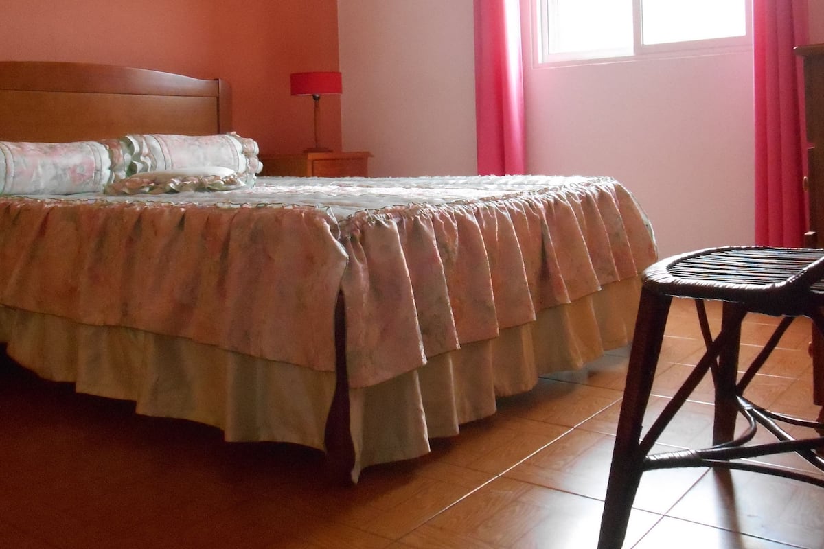 A bedroom with a bed, a chair, a nightstand, a lamp, and a window with curtains.
